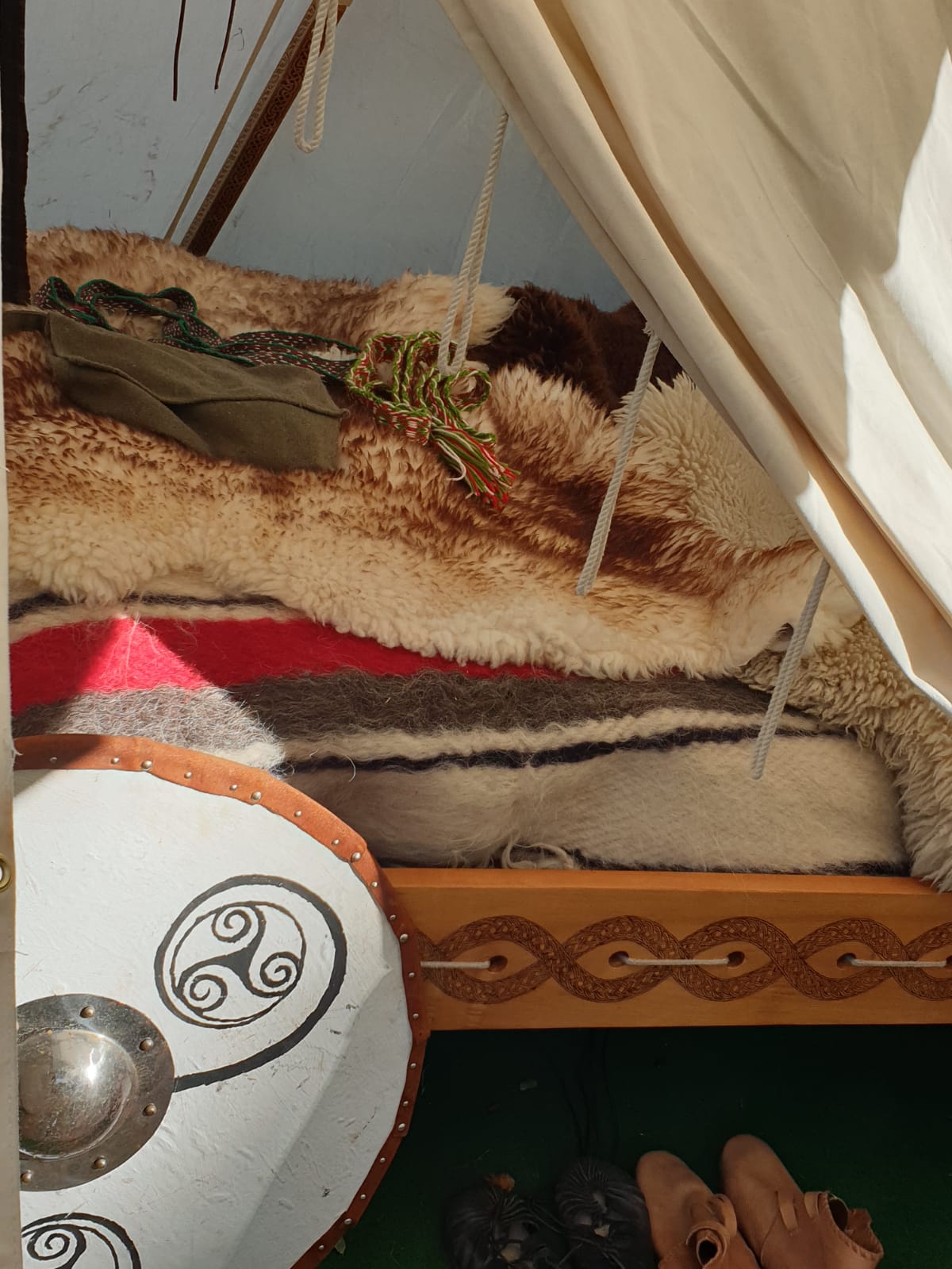 Unser Bett: Felle und Wolldecken auf geschnürtem Gestell, die Zarge mit Flechtband-Brandmalerei, davor ein Rundschild